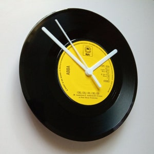 Puede incluir: Un reloj circular hecho de un disco de vinilo negro, con manecillas blancas. La esfera del reloj es una etiqueta amarilla con la palabra "ABBA" y el título de la canción "I DO, I DO, I DO, I DO, I DO".