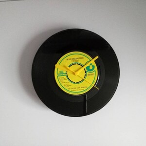 Puede incluir: Reloj circular hecho de un disco de vinilo negro. La esfera del reloj es amarilla con texto negro y manecillas amarillas. La etiqueta dice "In Old England Town" y "The Electric Light Orchestra". El reloj est&aacute; montado en una pared blanca.