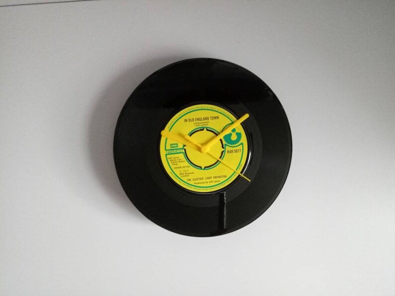 Puede incluir: Reloj circular hecho de un disco de vinilo negro. La esfera del reloj es amarilla con texto negro y manecillas amarillas. La etiqueta dice "In Old England Town" y "The Electric Light Orchestra". El reloj est&aacute; montado en una pared blanca.