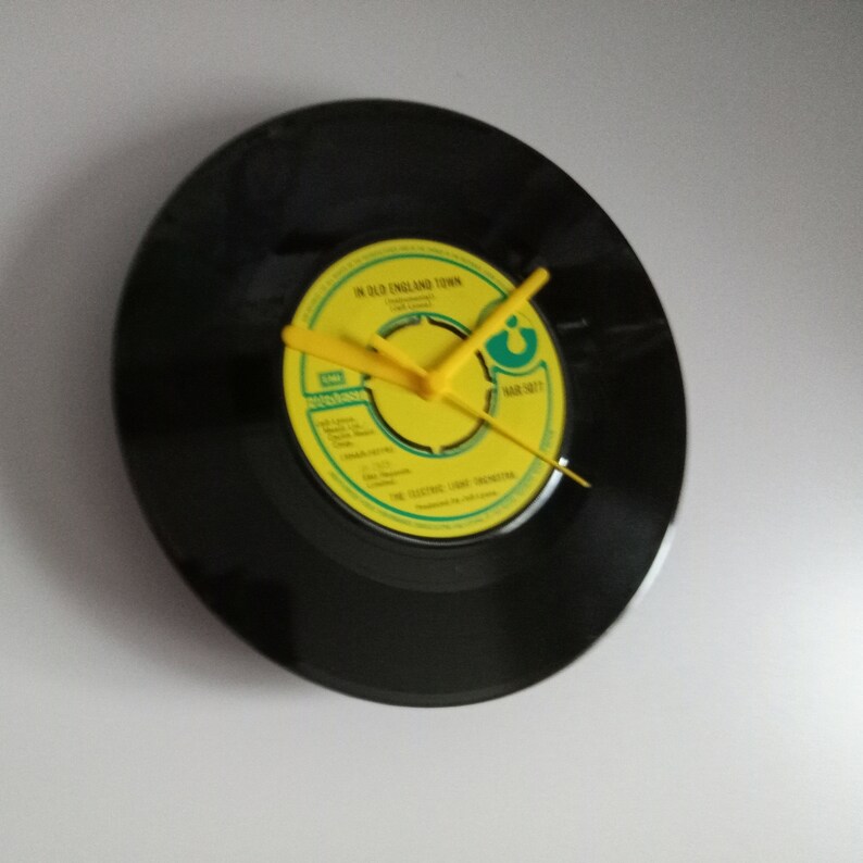 Puede incluir: Un reloj de vinilo negro con manecillas amarillas. La esfera del reloj es una etiqueta de disco amarilla con el texto "IN OLD ENGLAND TOWN" y "THE ELECTRIC LIGHT ORCHESTRA". Un reloj de pared &uacute;nico.