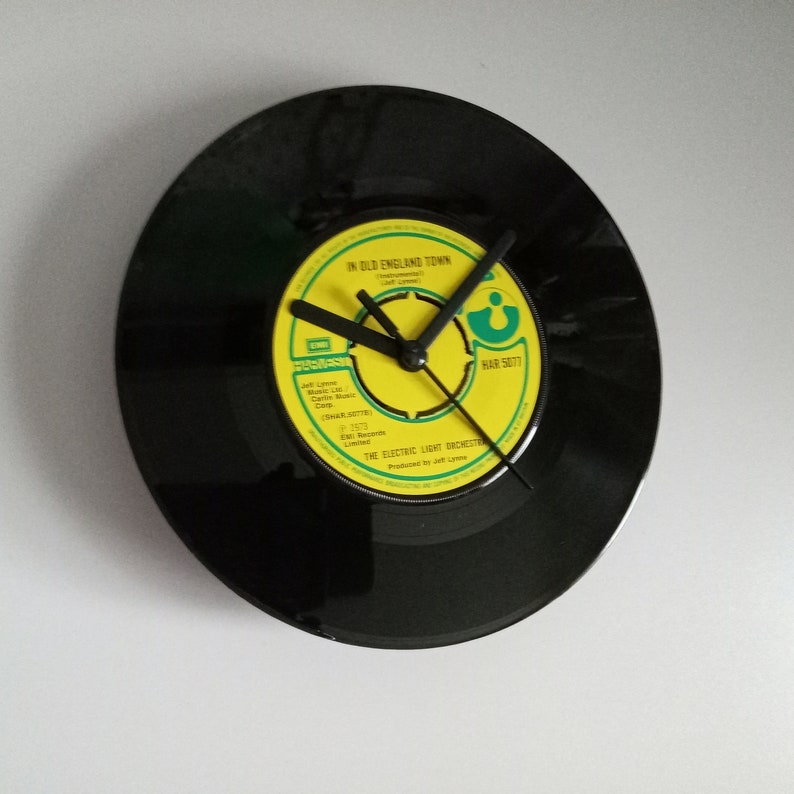 Puede incluir: Un reloj de vinilo negro con una etiqueta amarilla y verde. La etiqueta dice "IN OLD ENGLAND TOWN" y tiene manecillas negras. El reloj est&aacute; montado en una pared blanca.