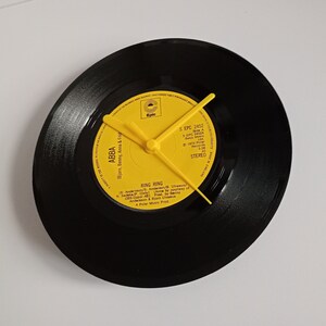Puede incluir: Un reloj de vinilo negro con manecillas amarillas y una etiqueta amarilla. La etiqueta dice "ABBA" y "RING RING". El reloj es un artículo decorativo único, perfecto para los amantes de la música.