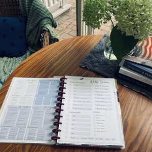May include: An open planner with a weekly schedule and daily theme sections. The planner is on a wooden table with a stack of books, including "Magnolia Table" and "We Made a Home".