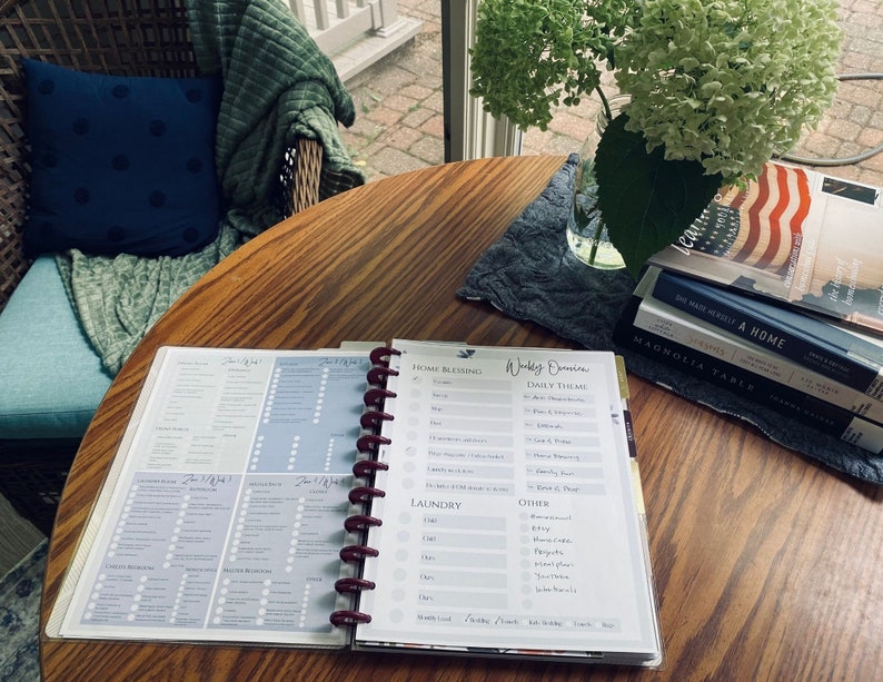 May include: An open planner with a weekly schedule and daily theme sections. The planner is on a wooden table with a stack of books, including "Magnolia Table" and "We Made a Home".