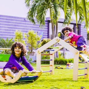 May include: Two children playing outdoors on wooden playground equipment. One child is on a rocking toy, and the other is climbing a ladder. The playground has a slide and colorful rungs. The logo "Thinkers Toys" is in the top left corner.