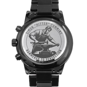May include: Black chronograph watch with a stainless steel band. The back of the watch features a silver engraved design of a man hammering a piece of metal on an anvil. The engraving reads "Simon "Hitter" Cummins Hammered to Perfection".