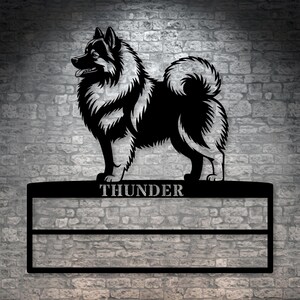 Op de afbeelding: Een wanddecoratie van zwart metaal met het silhouet van een pluizige hond boven een rechthoekig frame. De naam THUNDER is in hoofdletters uitgesneden in de bovenste balk van het frame, dat is voorzien van twee horizontale metalen rails.