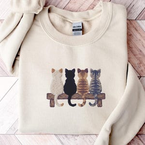 May include: A cream-colored sweatshirt featuring a graphic of four cats sitting on a wooden bench. The cats are in various colors: white, black, brown, and blue. The sweatshirt has a crew neck and long sleeves.