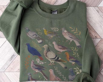 May include: An olive green crewneck sweatshirt featuring a graphic print of various pigeons and doves in shades of gray, blue, brown, and yellow, interspersed with small botanical leaf and flower illustrations across the front chest area.