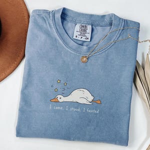 May include: A blue short-sleeved t-shirt with a graphic of a white duck lying down with yellow stars above its head. The text below the duck reads, I came. I stood. I fainted. The shirt has a Comfort Colors label in the neck area, size S-P-CH.