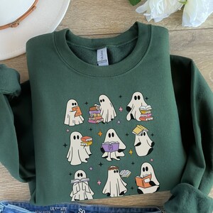 May include: A dark green sweatshirt featuring a design of several white ghosts reading books. The ghosts are depicted in different poses, some holding books, others with books on their heads. The sweatshirt has a crew neck and long sleeves.