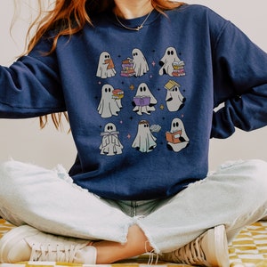 May include: A navy blue sweatshirt featuring a design of several ghosts reading books. The ghosts are white and have various expressions. The sweatshirt is being worn by a person sitting with their legs crossed.