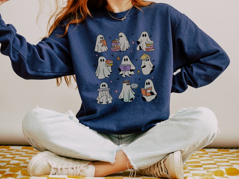 May include: A navy blue sweatshirt featuring a design of several ghosts reading books. The ghosts are white and have various expressions. The sweatshirt is being worn by a person sitting with their legs crossed.