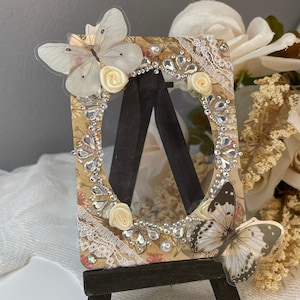May include: A small, ornate picture frame with a clear glass insert, decorated with white lace, faux pearls, and two butterflies. The frame is on a small wooden easel.
