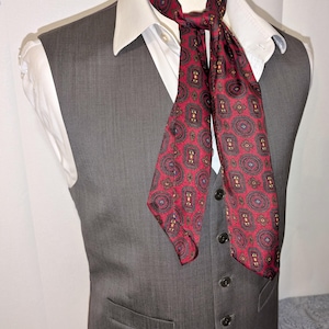May include: A grey waistcoat worn over a white collared shirt, with a patterned ascot. The ascot is red with a repeating paisley design in black, gold, and blue. The waistcoat has a button-down front.
