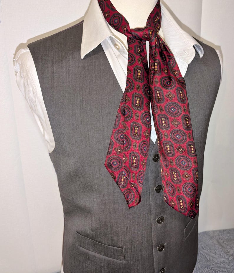 May include: A grey waistcoat worn over a white collared shirt, with a patterned ascot. The ascot is red with a repeating paisley design in black, gold, and blue. The waistcoat has a button-down front.