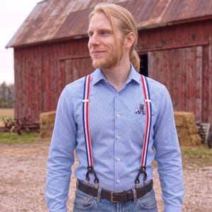 Può includere: Camicia azzurra con bretelle a righe rosse, bianche e blu. Le bretelle hanno attacchi in pelle e sono abbinate a una cintura marrone e jeans blu. Un fazzoletto da taschino fantasia è visibile nella tasca della camicia.