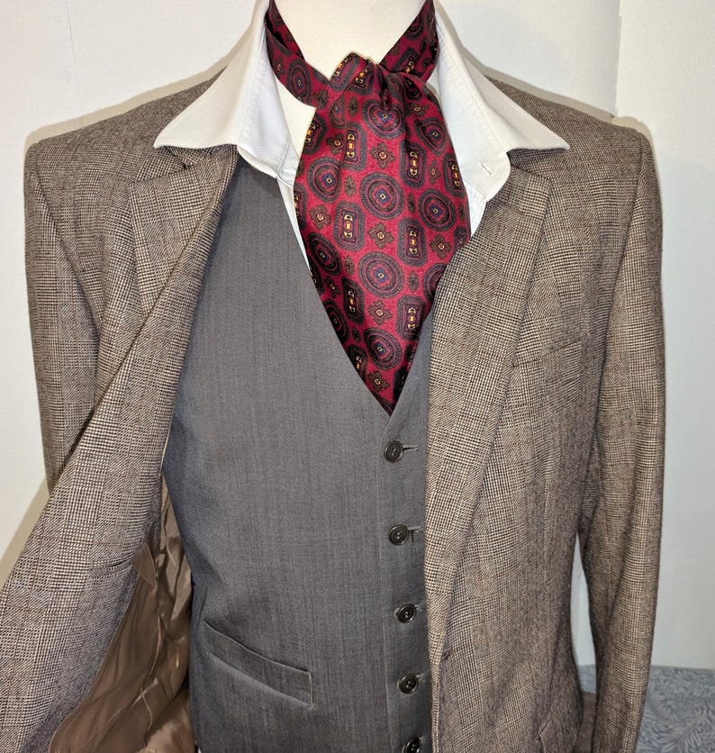 May include: A brown check blazer, grey waistcoat, and burgundy paisley ascot. The blazer is worn over a white collared shirt. The waistcoat has a button-up front. The ascot is knotted at the neck, creating a formal look.