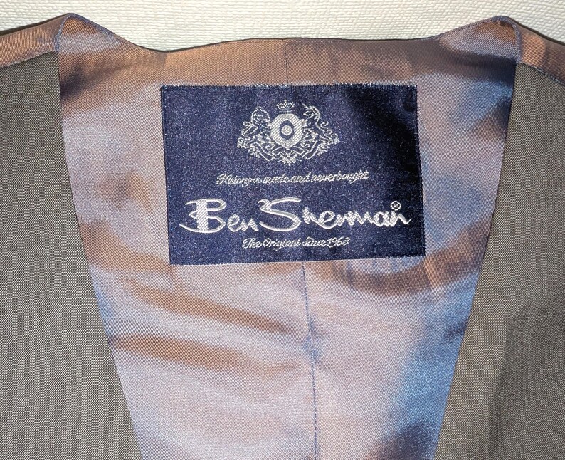 May include: Close-up of the inside of a grey waistcoat. The lining is a pale blue-grey colour. A navy blue label with the Ben Sherman logo and text is sewn into the lining. The label also includes the text "The Original Since 1963."