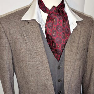 May include: A brown plaid blazer with a grey waistcoat and a white collared shirt. A red patterned ascot is knotted at the neck. The suit is displayed on a mannequin.
