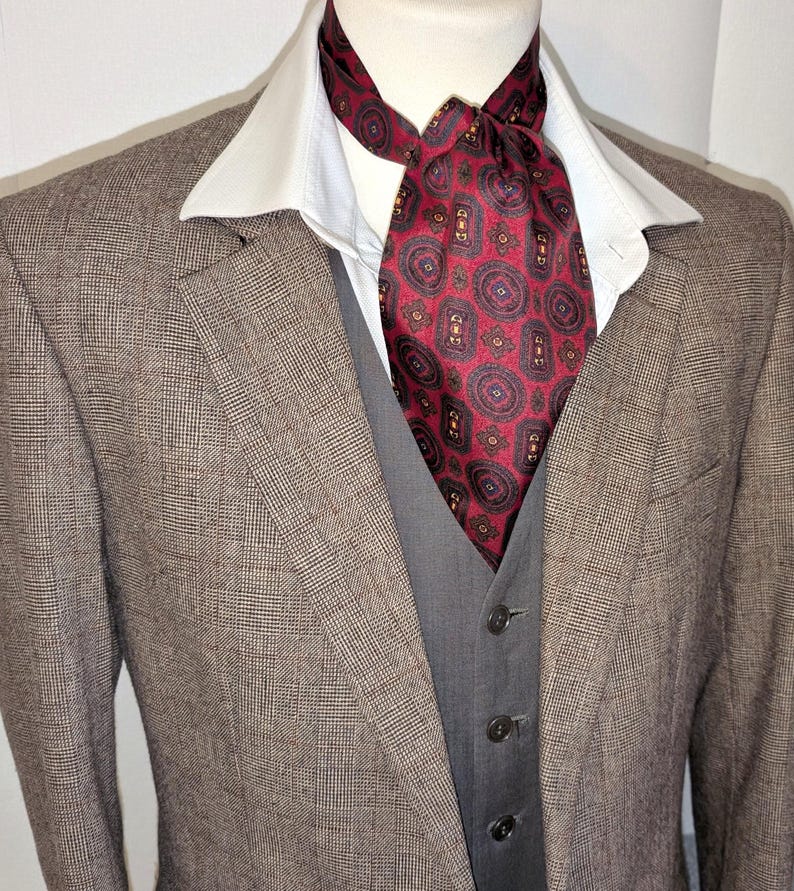 May include: A brown plaid blazer with a grey waistcoat and a white collared shirt. A red patterned ascot is knotted at the neck. The suit is displayed on a mannequin.