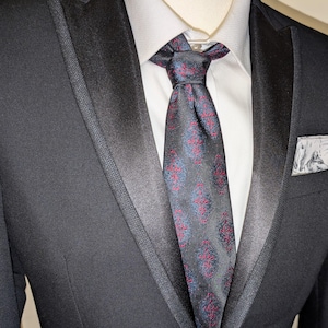 May include: A black tuxedo jacket with a white dress shirt and a patterned tie. The tie has a dark background with red and blue floral designs. A pocket square with a grey and white design is in the breast pocket.