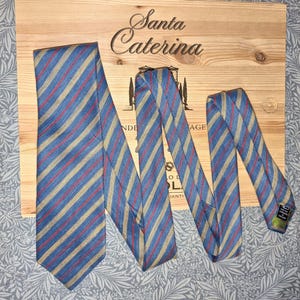 May include: A blue tie with red and tan stripes is displayed on a wooden surface. The tie is made of a woven fabric and has a classic design. The background is a wooden surface with the words "Santa Caterina" on it.