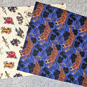 Könnte beinhalten: Zwei Stoffmuster. Eines ist cremefarben mit Tattoo-Illustrationen von Rosen, Dolchen, Totenköpfen und Adlern. Das andere ist blau mit einem sich wiederholenden Muster aus orangen Tigern und schwarzen Blättern.