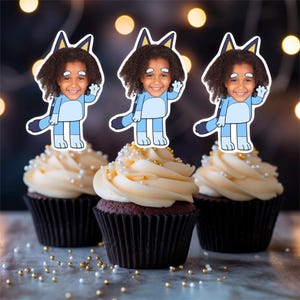 May include: Three chocolate cupcakes topped with vanilla frosting and edible gold and silver sprinkles. Each cupcake is decorated with a cartoon cutout of a child in a blue costume, waving.