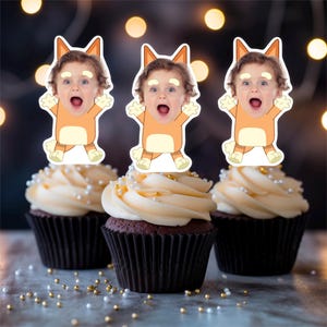 May include: Three chocolate cupcakes with white frosting, topped with cartoon character cutouts. The cupcakes are decorated with gold and silver sprinkles. The background is blurred with bokeh lights.