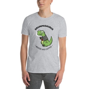 Puede incluir: Una camiseta gris con una ilustración de dibujos animados de un dinosaurio verde con traje y sosteniendo una taza de café. El texto "Grumpasaurus - Needs His Coffee -" está impreso en la camiseta.