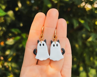 May include: A pair of dangle earrings featuring white ghost-shaped charms with black cat ears and tails. The earrings are made of polymer clay and have gold-colored hooks.