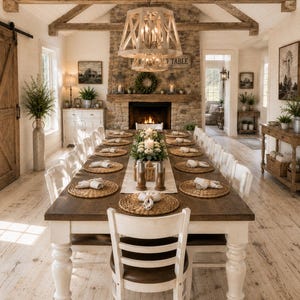 May include: A long rectangular dining table with a dark wood top and white turned legs, surrounded by white chairs with dark seats. The table is set with woven round placemats, white napkins, and a central floral arrangement with two candles.