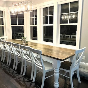 May include: A long, wooden farmhouse table with white legs and a white top. The table is set with white chairs and is positioned in front of a window.