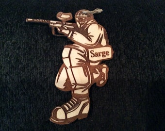 May include: A wooden cutout of a person holding a paintball gun. The cutout is brown and has a name tag that says "Sarge".