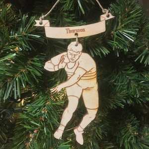 May include: A wooden Christmas ornament depicting a shot put athlete in action. The ornament is light brown and has the name "Thorsson" engraved on a banner above the figure. The ornament is hanging on a green Christmas tree.