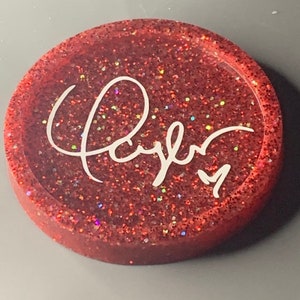 K&ouml;nnte beinhalten: Ein roter Glitzeruntersetzer mit dem Wort "Kayla" in wei&szlig;er Kursivschrift.