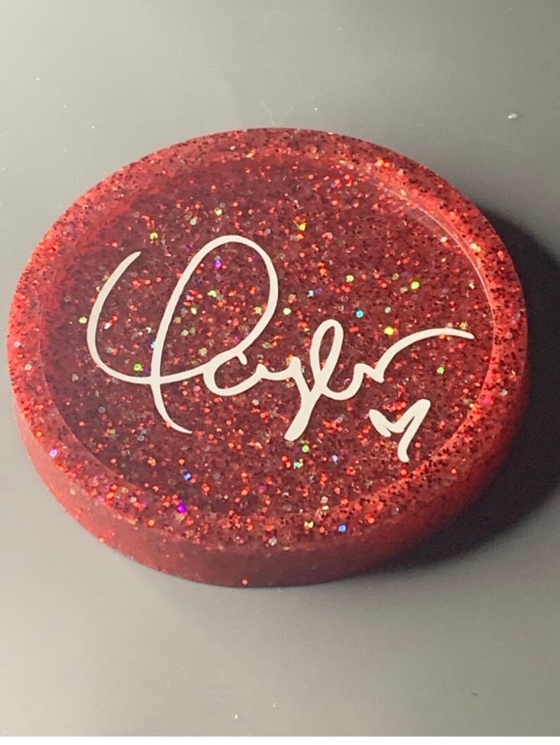 K&ouml;nnte beinhalten: Ein roter Glitzeruntersetzer mit dem Wort "Kayla" in wei&szlig;er Kursivschrift.