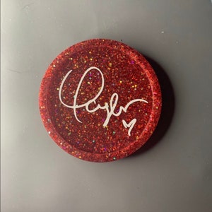 K&ouml;nnte beinhalten: Roter Glitzer-Untersetzer mit einer wei&szlig;en, kursiv geschriebenen Signatur, die "Taylor" lautet.