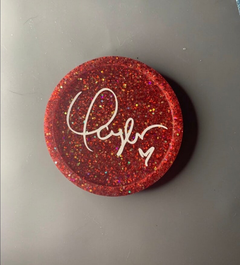 K&ouml;nnte beinhalten: Roter Glitzer-Untersetzer mit einer wei&szlig;en, kursiv geschriebenen Signatur, die "Taylor" lautet.