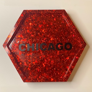 Puede incluir: Posavasos hexagonal rojo brillante con la palabra "CHICAGO" en letras negras. El posavasos está hecho de resina y tiene un acabado brillante. El brillo es una mezcla de rojo y naranja, creando un efecto brillante.
