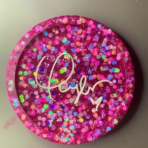 K&ouml;nnte beinhalten: Runder rosa Untersetzer mit glitzernder Oberfl&auml;che und dem Namen "Taylor" in wei&szlig;er Kursivschrift.