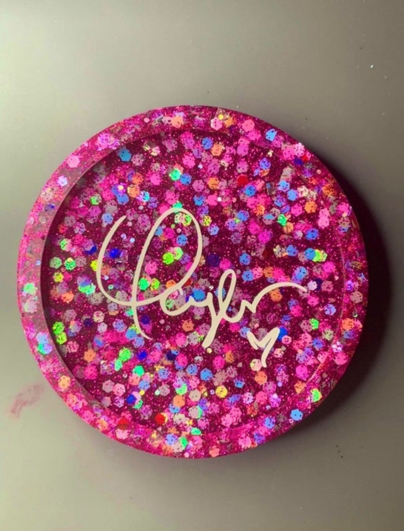 K&ouml;nnte beinhalten: Runder rosa Untersetzer mit glitzernder Oberfl&auml;che und dem Namen "Taylor" in wei&szlig;er Kursivschrift.