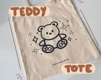 May include: A beige canvas tote bag with black and white illustrations of a teddy bear and stars. The bag has brown text that says "TEDDY" and "TOTE".
