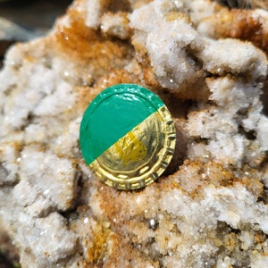 May include: A green and gold bottle cap with a diagonal color split. The green section is on the top left, and the gold section has a logo. The cap is resting on a rough, textured, light-colored rock surface.
