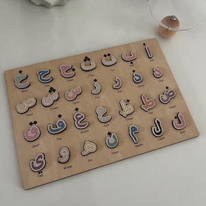 Könnte beinhalten: Holzpuzzle mit dem arabischen Alphabet. Jeder Buchstabe ist ein separates Stück, bemalt in Pastellfarben und mit seinem Namen beschriftet. Das Brett ist aus hellem Holz gefertigt.