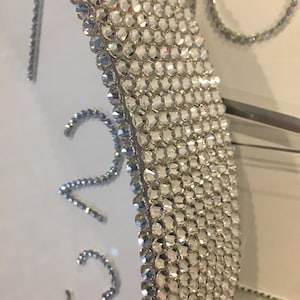 May include: Close-up of a decorative wall clock with a white face and silver rhinestone embellishments. The clock features silver numerals and a sparkling, crystal-covered border. The design is elegant and glamorous.