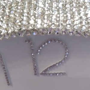May include: Close-up of a white clock face with the number 12 prominently displayed, embellished with sparkling crystals. The clock's edge is also adorned with crystals, creating a glamorous, decorative timepiece.