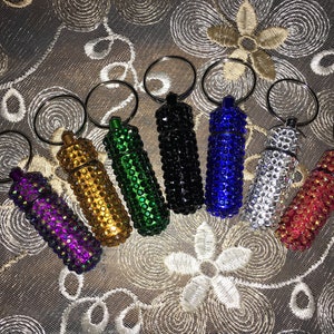 May include: An assortment of seven small, cylindrical containers, each encrusted with sparkling rhinestones in various colors: purple, gold, green, black, blue, silver, and red. Each has a silver keyring attached.