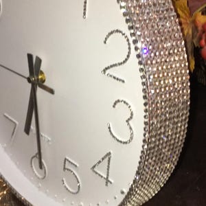 May include: White wall clock with silver rhinestone accents around the edge and on the numbers. The clock hands are black, and the numbers are in a simple, elegant font. The clock face is white.